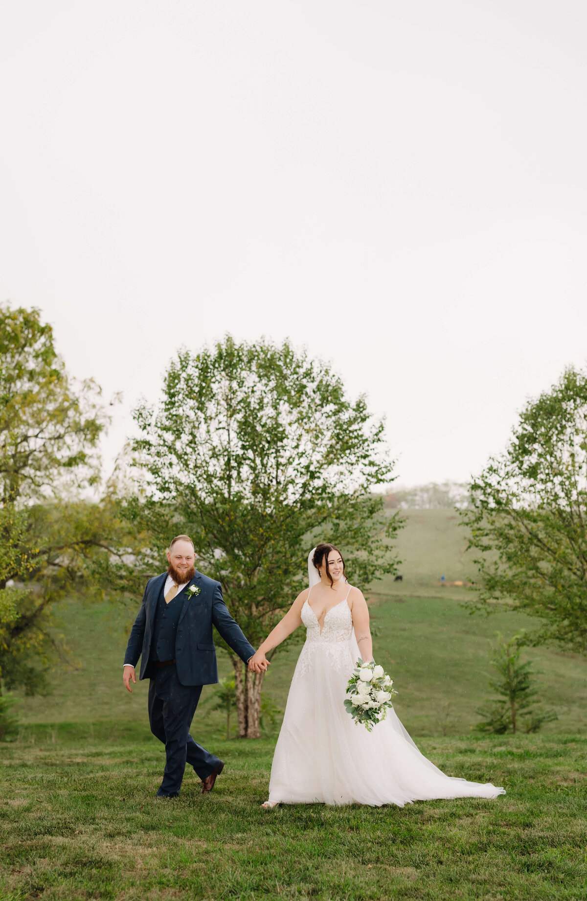 groom holding his brides hand and leading her through a field at Sunny Slope Farm as they weave through trees together on the farm for a spring wedding photographed by Charlottesville wedding photographer