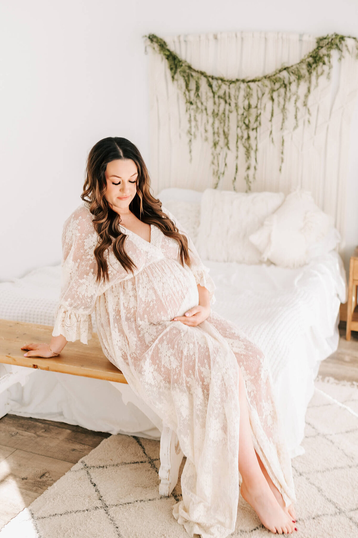 pregnant mom in a lace dress looking down at her baby bump while sitting on a bench in studio captured by Springfield MO maternity photographer Jessica Kennedy of The XO Photography