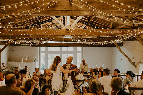 Mariée dans avec son papa sous les regards des invités dans la salle de réception surmontée de poutres en bois et des guirlandes de lumière. Photo capturée par Laura, photographe de mariage en Vendée.