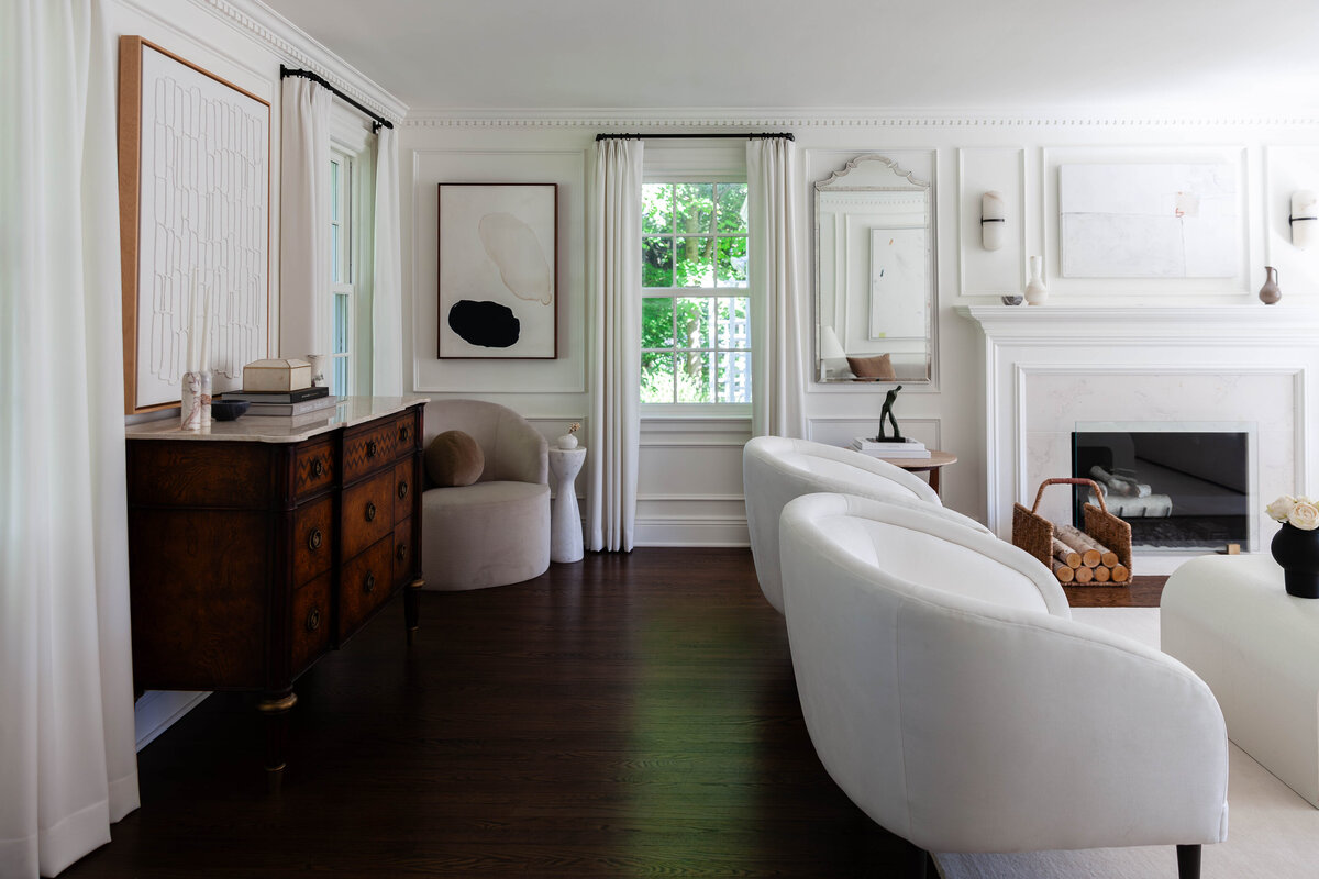 This elegantly curated living room showcases a harmonious blend of modern sophistication and timeless charm. Featuring plush white accent chairs, an ornate antique wood sideboard, and layered neutral textures, this space exudes a refined aesthetic. The custom millwork, curated artwork, and a sleek marble fireplace add an extra touch of luxury interior design. Thoughtfully selected décor pieces, including sculptural vases and soft textiles, enhance the warm yet sophisticated ambiance.  Perfect for homeowners in Short Hills, Summit, Montclair, and Maplewood, this high-end interior design reflects a polished yet inviting aesthetic. If you're seeking luxury interior design services in New Jersey, we specialize in crafting elevated, bespoke spaces that blend beauty with functionality.