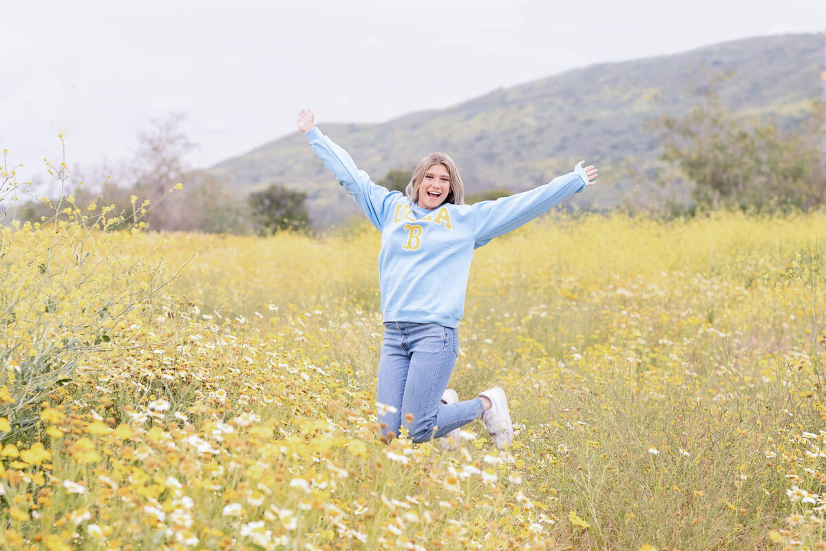A person wearing a light blue sweatshirt and jeans joyfully kneels in a field of yellow and white flowers, arms spread wide. A hill and cloudy sky are in the background.