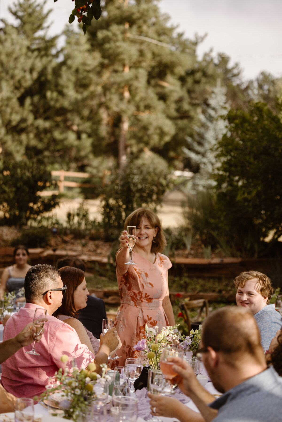 A woman in a floral dress stands smiling and raising a glass for a toast at an outdoor wedding reception surrounded by guests at a long table.
