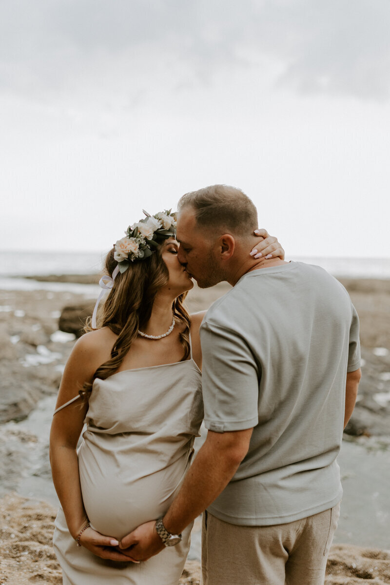 Femme enceinte de face embrasse son mari à sa droite de dos. Il lui caresse son ventre rond. Shooting photo grossesse réalisé à la plage en vendée.