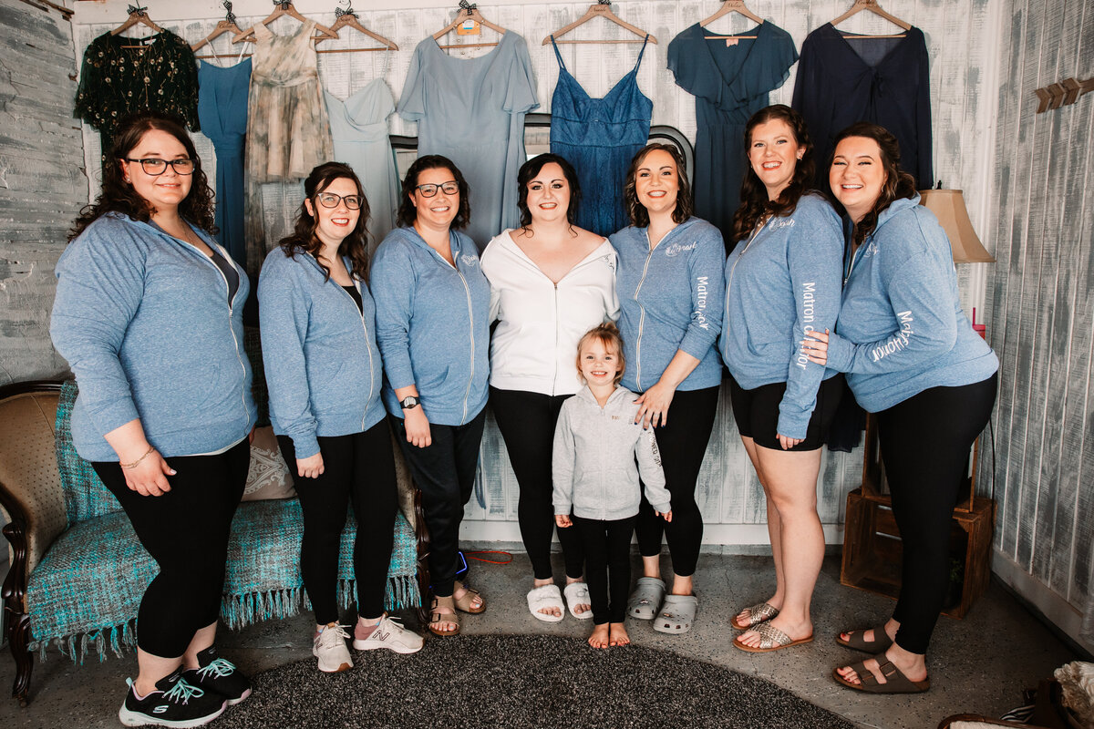 A bride stands in a rustic room with her 6 bridesmaids and flower girl in matching hoodies