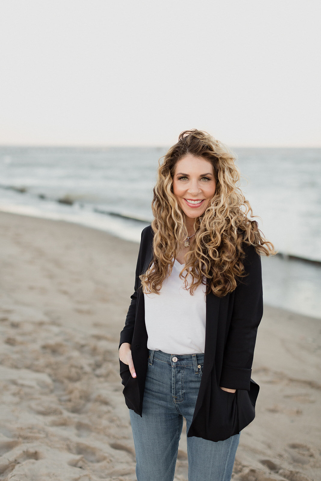 woman is on beach, portrait of her looking at camera and smiling. She is wearing a black cardigan, white tee and jeans with her hands in her sweater pockets.