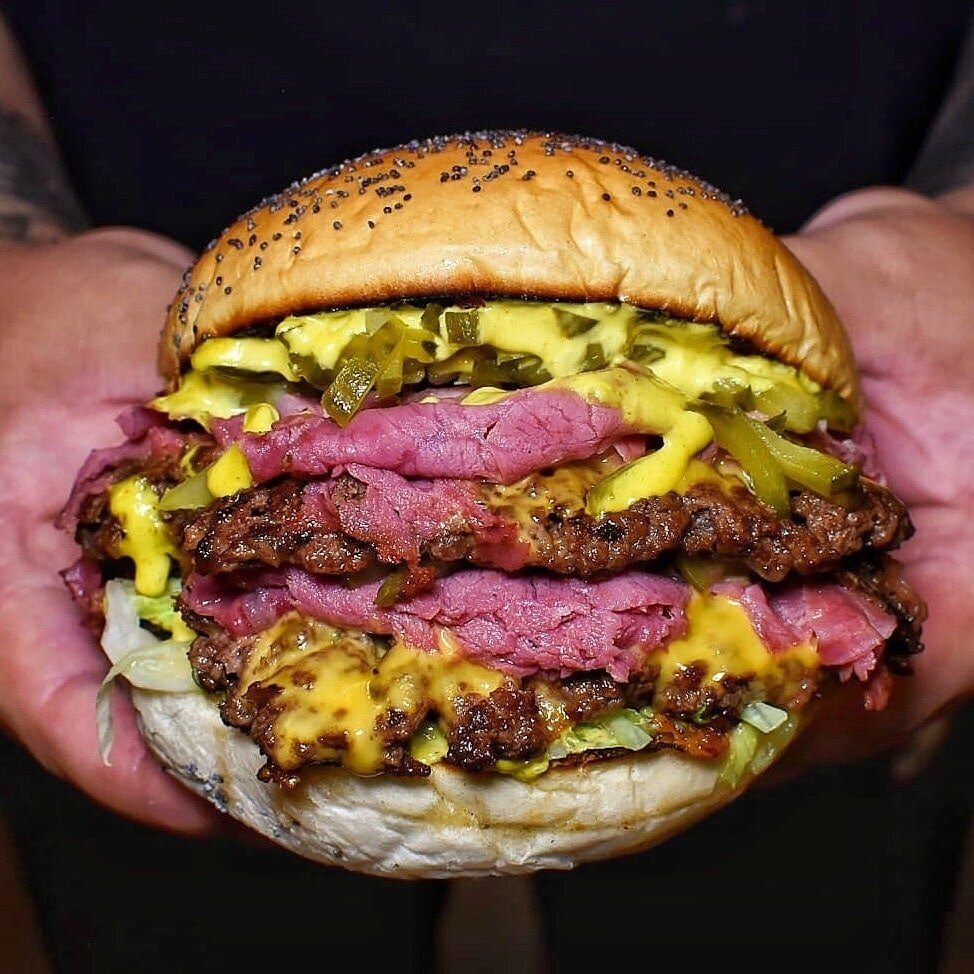 hands holding double beef burger, which is huge and stuffed full of pastrami, cheese and gherkins