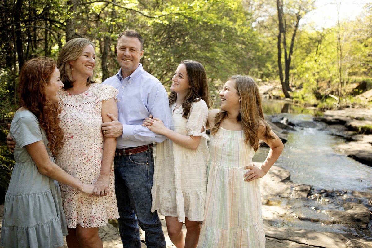 Three sisters smiling together by the creek, with their parents nearby, capturing memories during a family photo session