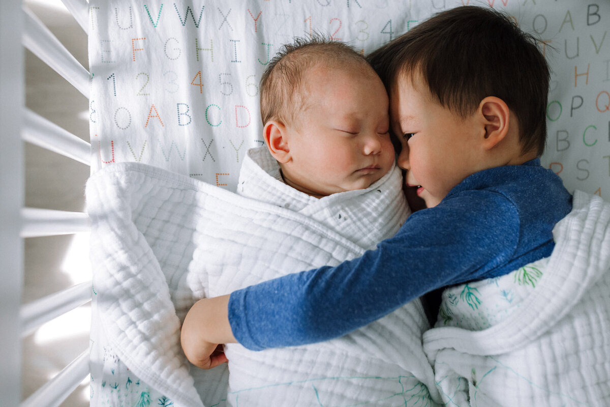 brother snuggling swaddled newborn brother in crib with both wrapped in white blanket