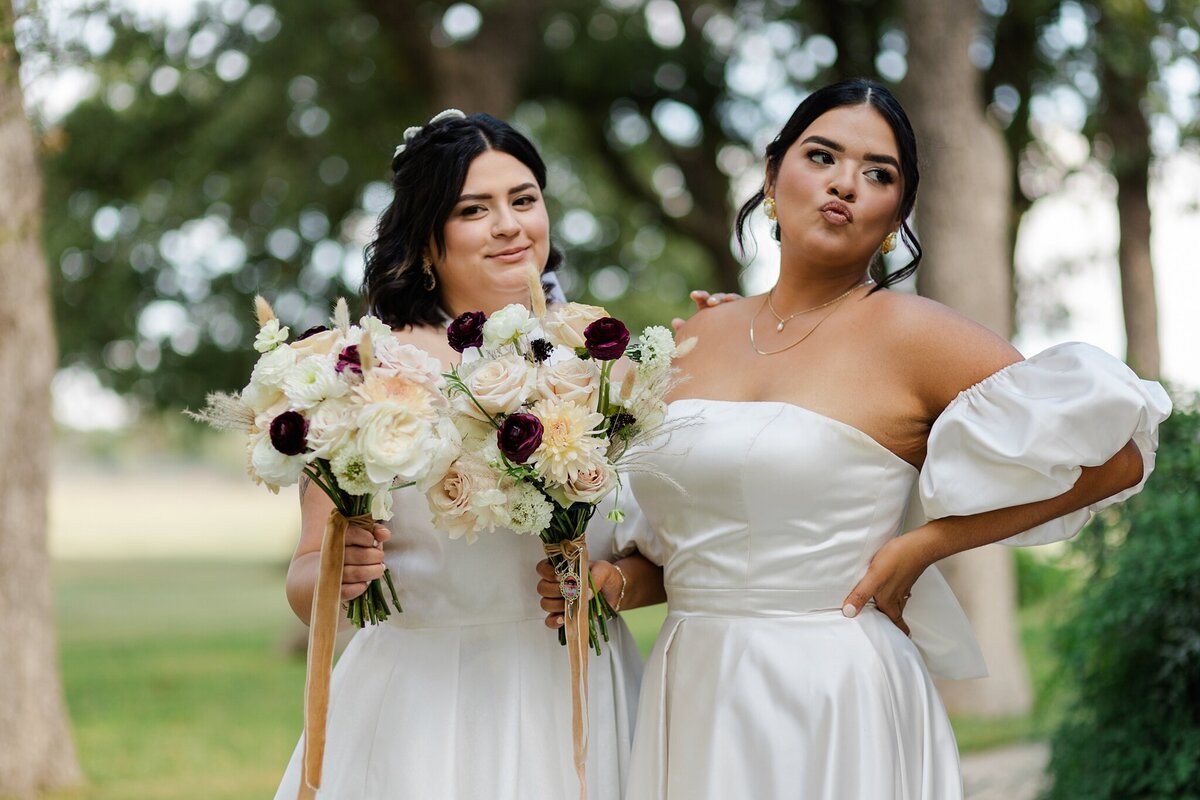 A candid shot of two brides during their wedding day at a wedding in Fort Worth, Texas. The bride on the left is looking right at the camera, is wearing a white dress, and is holding a bouquet. The bride on the right is unaware of the camera and is looking off to her left. She's wearing a white dress with large sleeves, and is holding a bouquet.