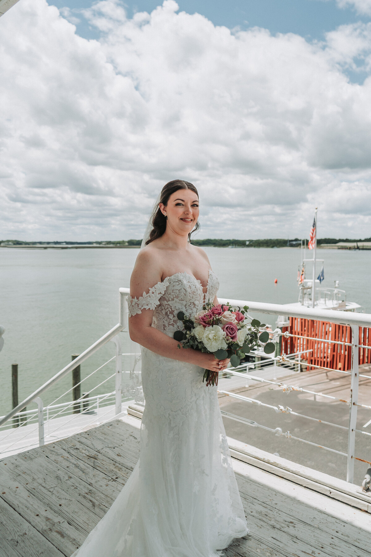 A beautiful bride in an off-the-shoulder lace wedding gown holds a colorful bouquet, smiling on a wooden balcony overlooking the water at the Lesner Inn in Virginia Beach, captured by Quinn Photography LLC.
