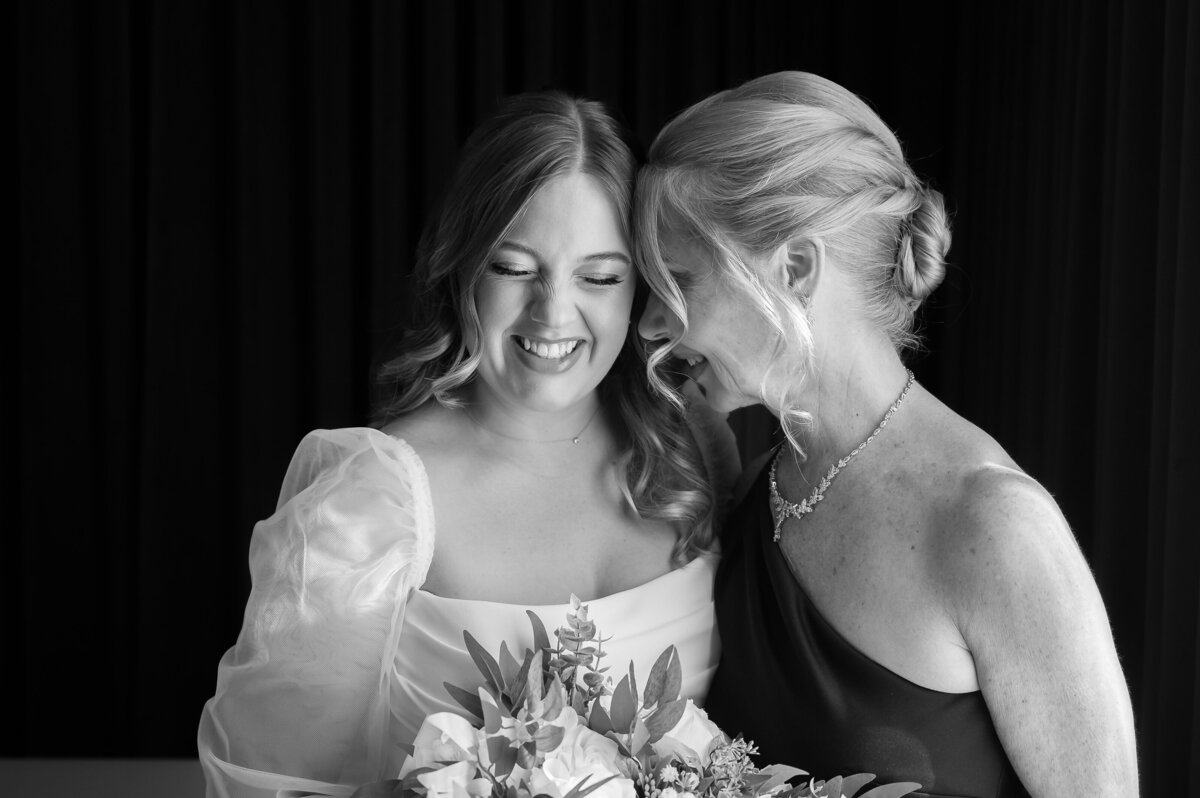 Two women stand close, smiling warmly in a classic wedding photography moment. One holds a bouquet and wears a dress with puffed sleeves; the other dons an elegant sleeveless dress. Behind them, a dark curtain adds depth to this Philadelphia wedding scene captured by their talented Philly wedding photographer.