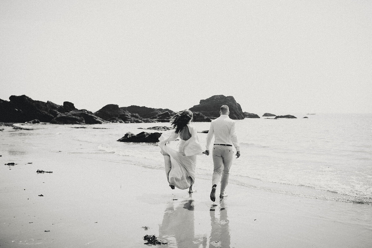 Bride and groom walking barefoot along the shoreline during their Maine wedding, captured in black and white.