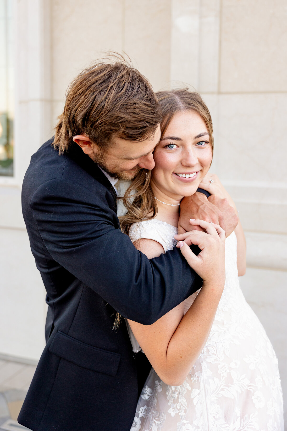 A newlywed couple embraces joyfully, with the groom resting his head on the bride's shoulder as she smiles at the camera