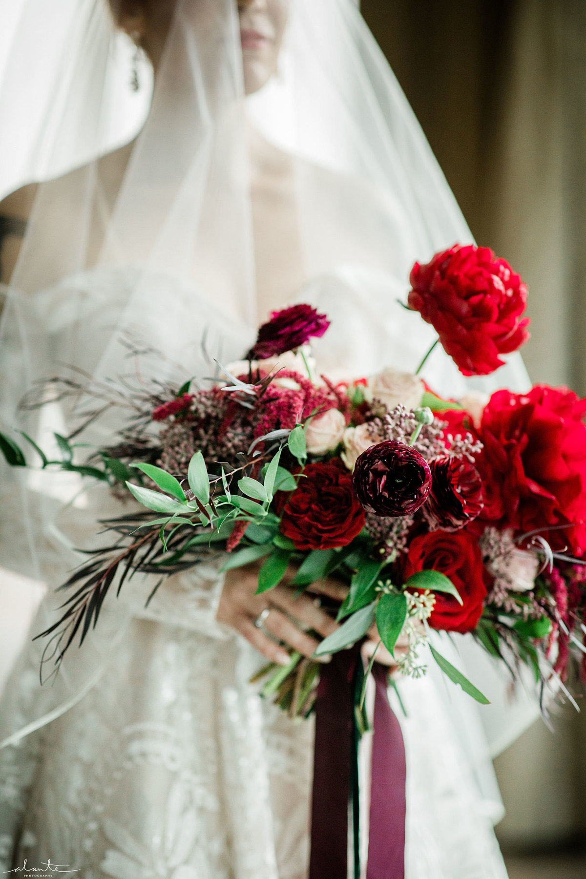 Deep red winter wedding bouquet of burgundy peonies, deep red roses, merlot ranunculus, greenery, and trailing silk ribbon streamers