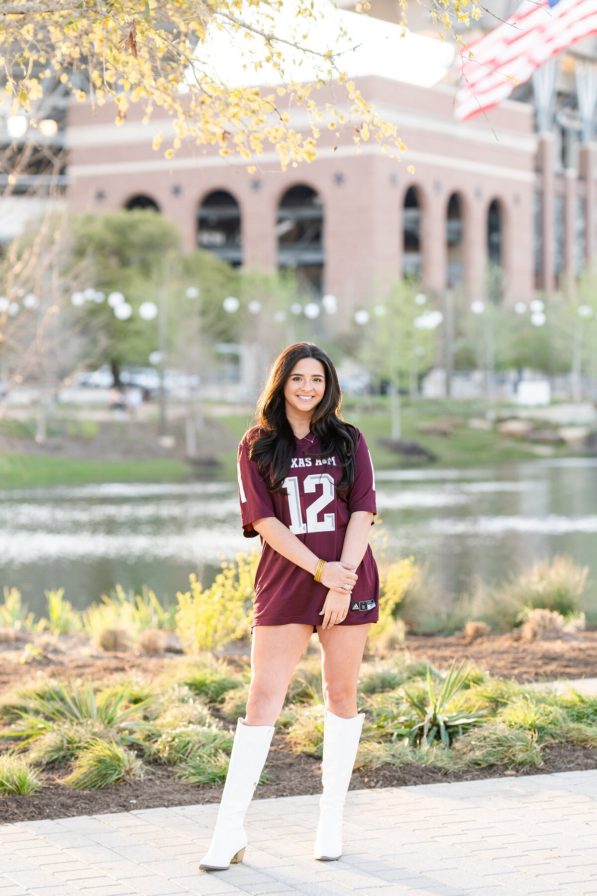 Texas A&M senior girl standing with hand on wrist and smiling in maroon jersey and white boots in Aggie Park in front of Kyle Field