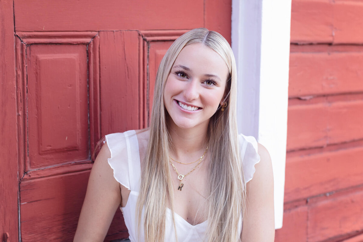 A person with long blonde hair is smiling in front of a red wooden door. They are wearing a white top and a gold necklace.