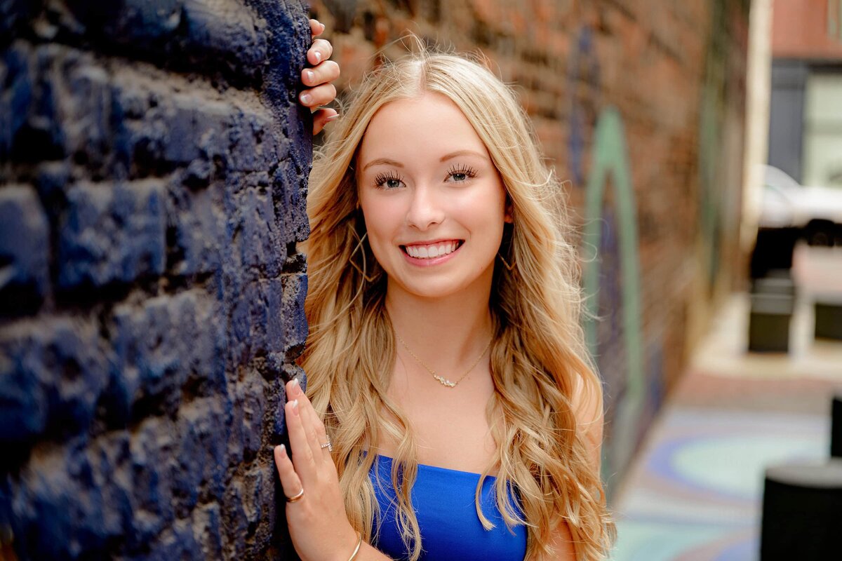 cuthbertson senior nikki stands next to a wall in rock hill, sc with her blonde hair falling down her shoulders and wearing a blue dress during her senior photo session