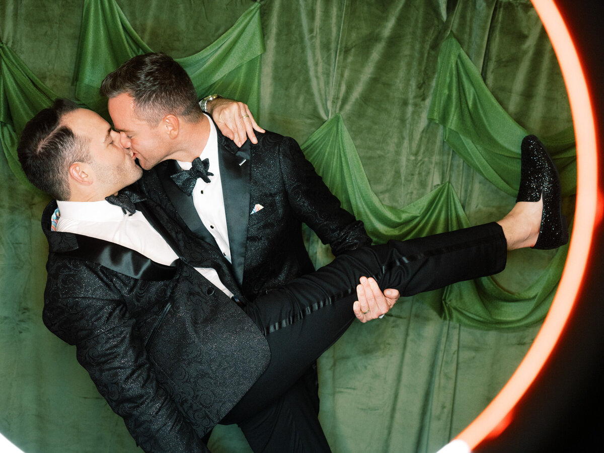 A joyful couple dressed in stylish suits share a loving kiss against a vibrant green backdrop, capturing a moment of deep affection and celebration of love.