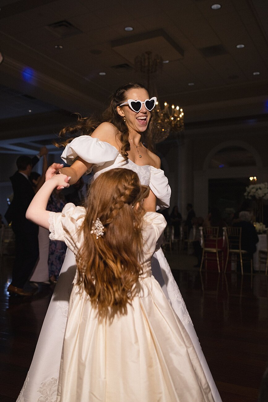 Bride wearing hear-shaped shades while dancing with kid at reception