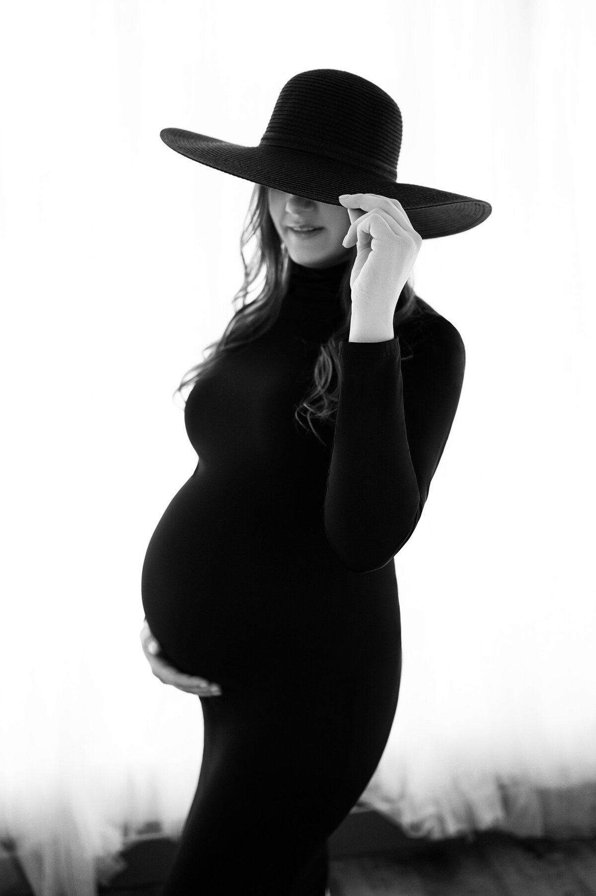 Black and white image of a pregnant woman in a black bodycon maternity dress and black sunhat that covers her eyes.
