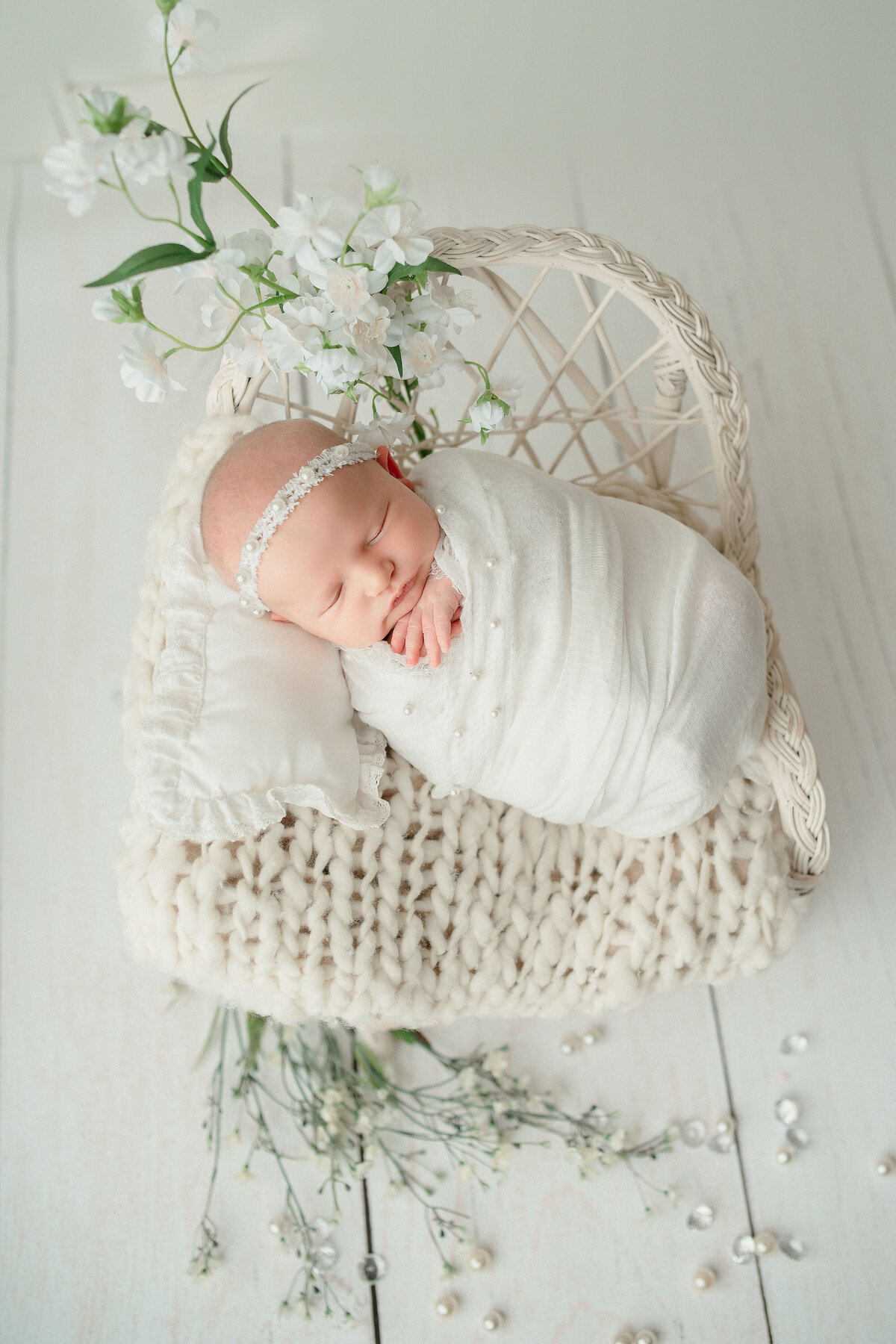Sleeping 2 week old baby girl in white swaddle laying head on tiny white pillow propped up in white bohemian  miniature chair