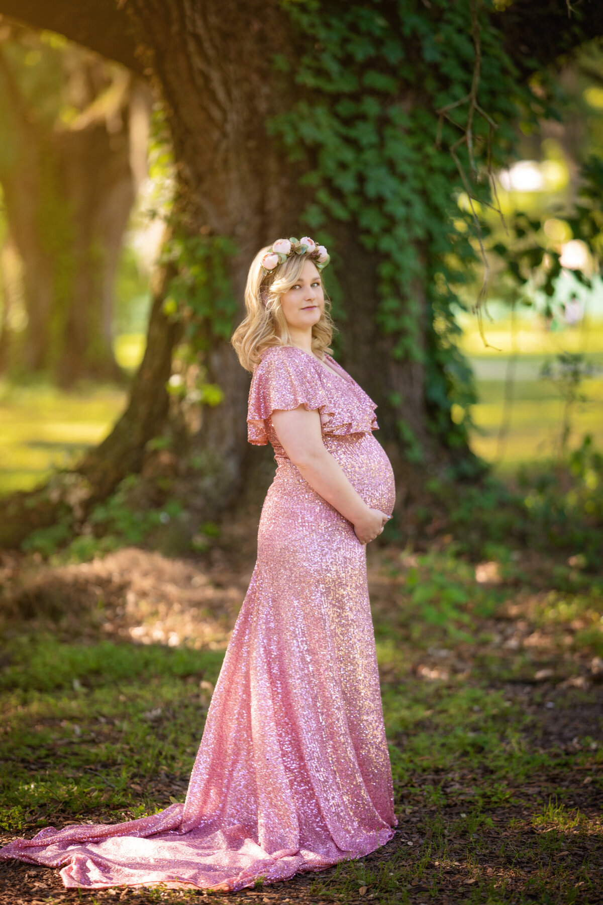 Blond woman with shoulder length hair and a pink floral crown stands in Audubon Park under a live oak tree.  She is pregnant with twins and is holding her belly.  She is wearing a pink sequined dress by Mii-Estillo with a long long train behind her.