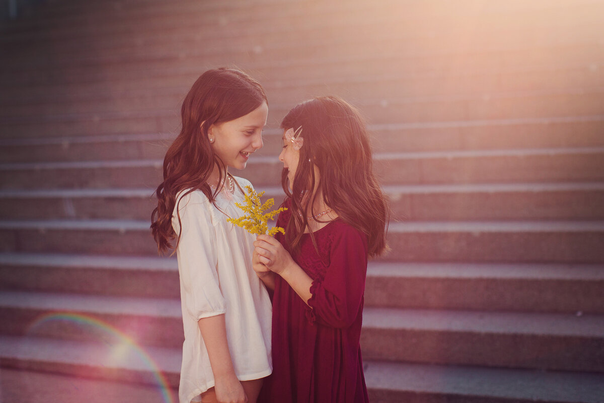 Strong light is coming in between two girls in a red dress and white dress, holding a yellow flower.