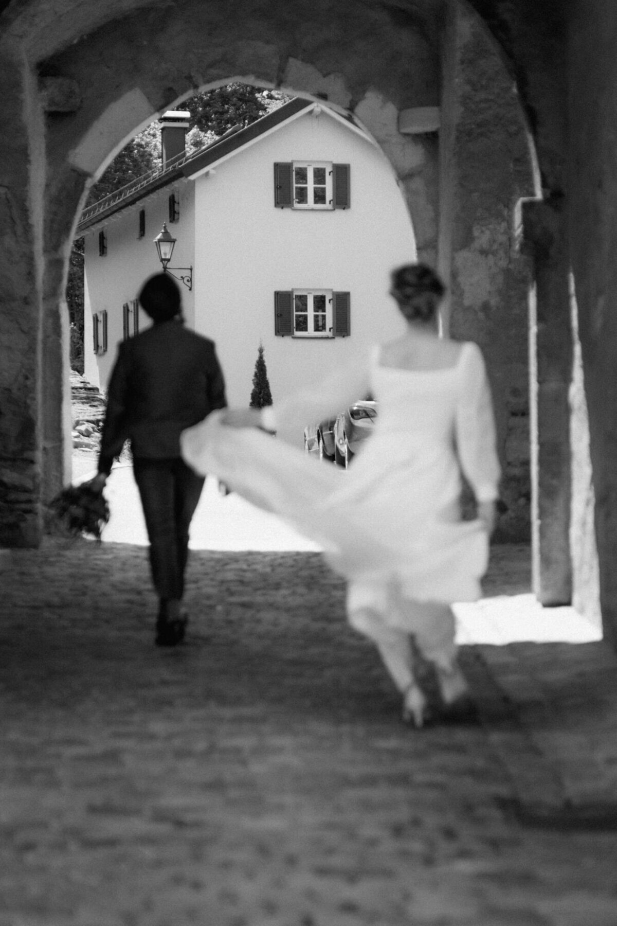 In einer Gasse mit einem Steinbogen läuft die Braut dem Bräutigam mit schwingendem Kleid nach.