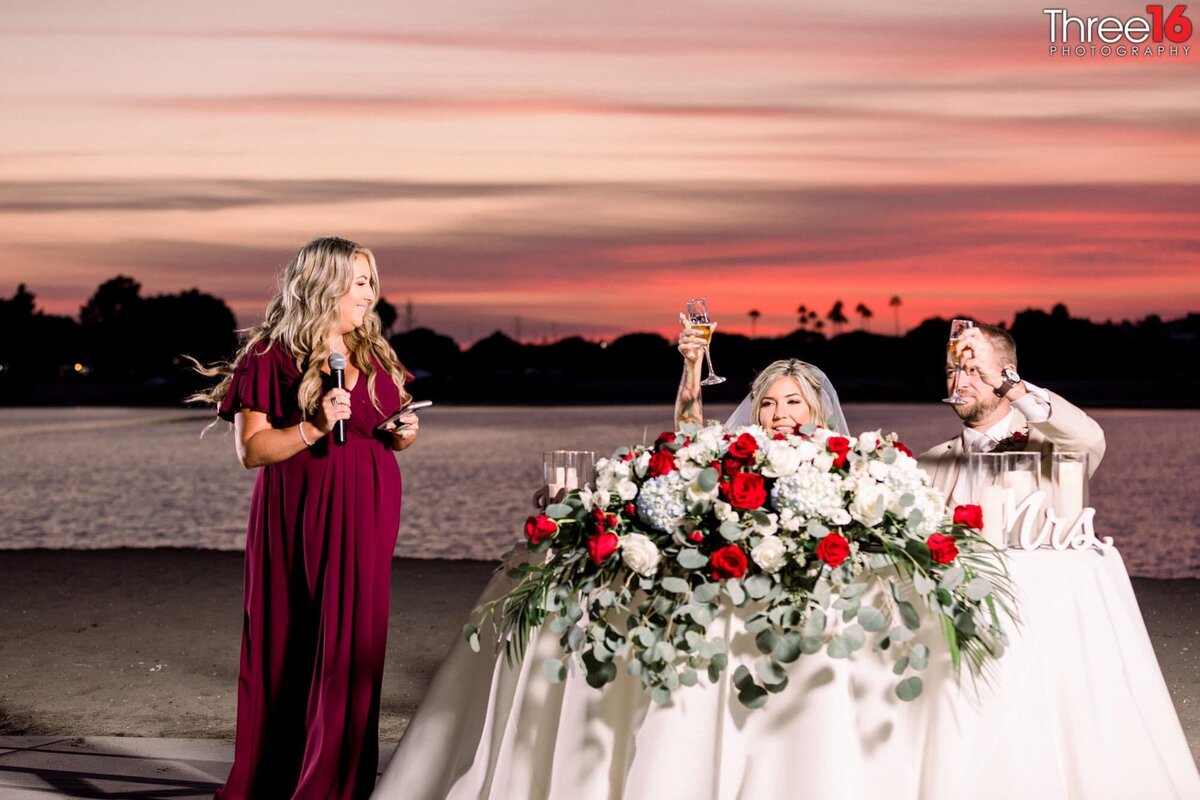 Maid of Honor toasts the couple as they sit at the sweetheart table behind a beautiful floral arrangement lifting their toasting glasses