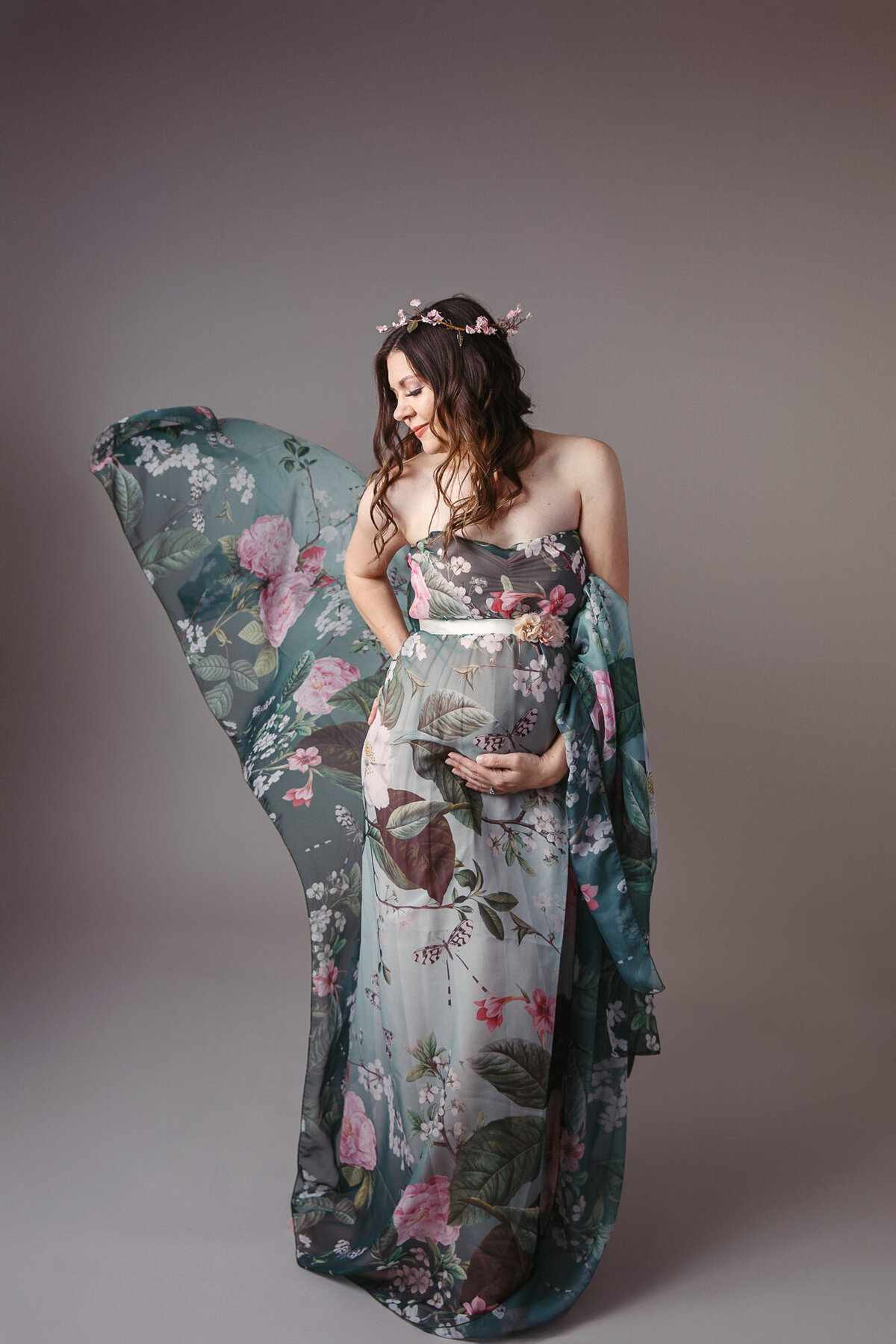 Beautiful woman holding her pregnant belly wearing a green floral gown that is blowing in the air behind her