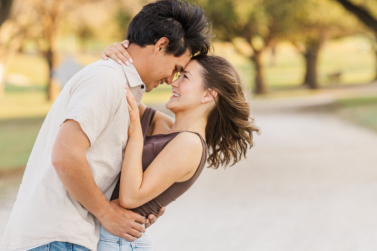 Candid photo of a joyful couple during their Fort Worth engagement session, sharing a sweet moment on a tree-lined path. Their connection and laughter reflect a playful, romantic energy perfect for save-the-date inspiration.