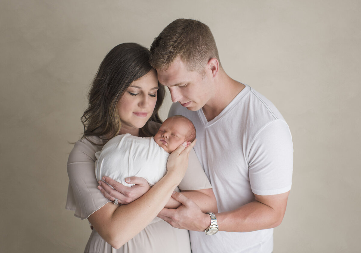 a mother and father with their newborn baby and a tan backdrop behind them