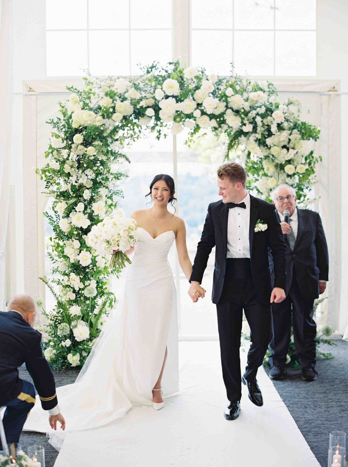 bride and groom smiling after the wedding ceremony as they walk down the aisle with white and green floral arch behind them