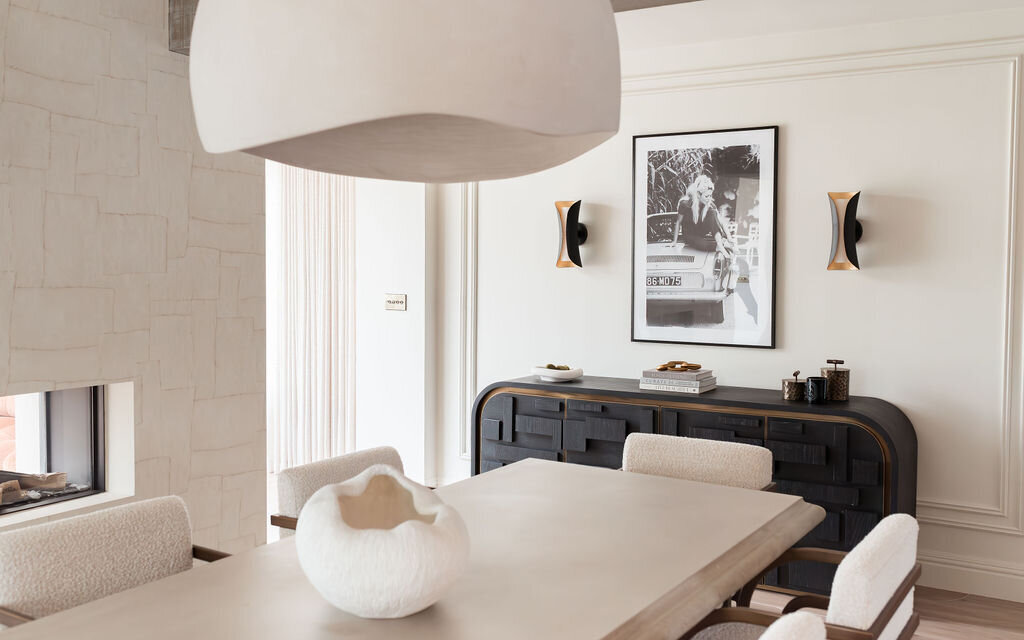 A modern dining room features a large light-colored table with beige padded chairs. On the wall, a black sideboard with a photograph above it is flanked by two contemporary sconces. The space is minimalistic with neutral tones and a sculptural white centerpiece on the table.