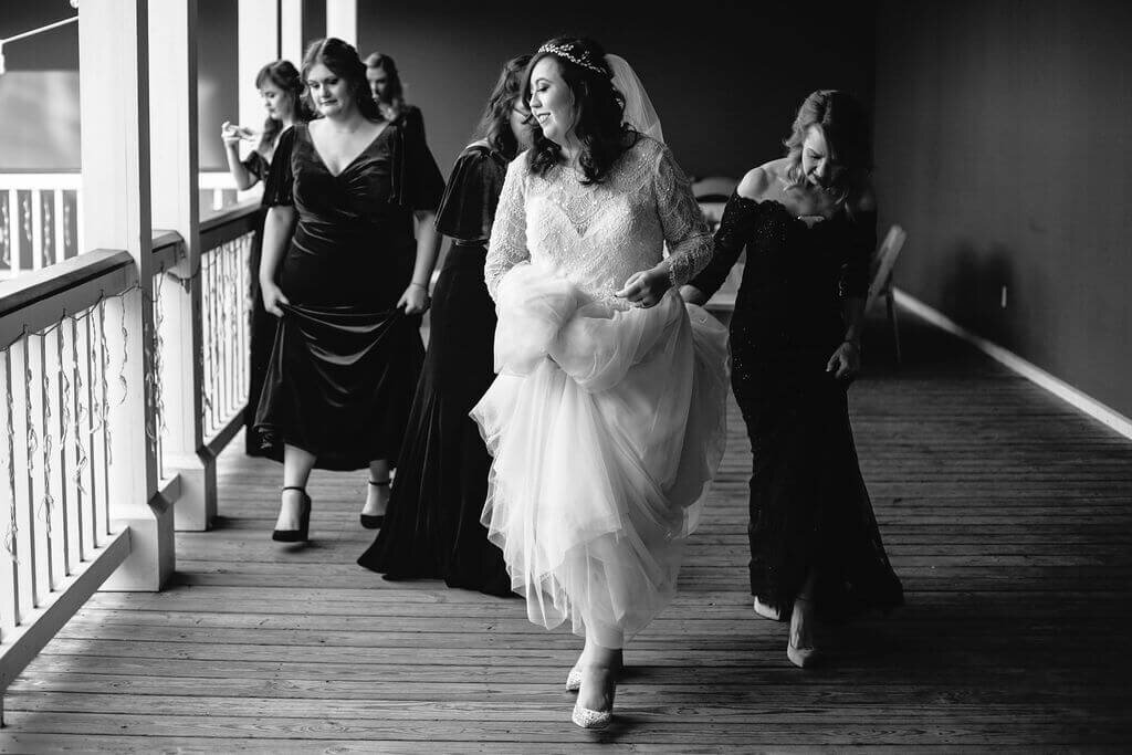 A bride in a white dress walks happily down a wooden hallway, holding her gown, followed by bridesmaids in dark dresses. The scene is joyful and candid, with natural light streaming in from the left.