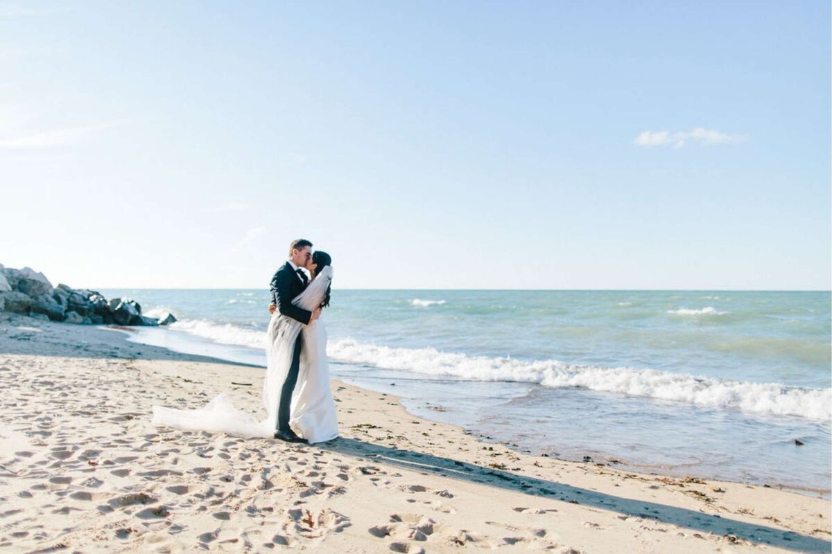 Bride and Groom Embrace during Portraits along the Beach for a Luxury Michigan Lakefront Golf Club Wedding.