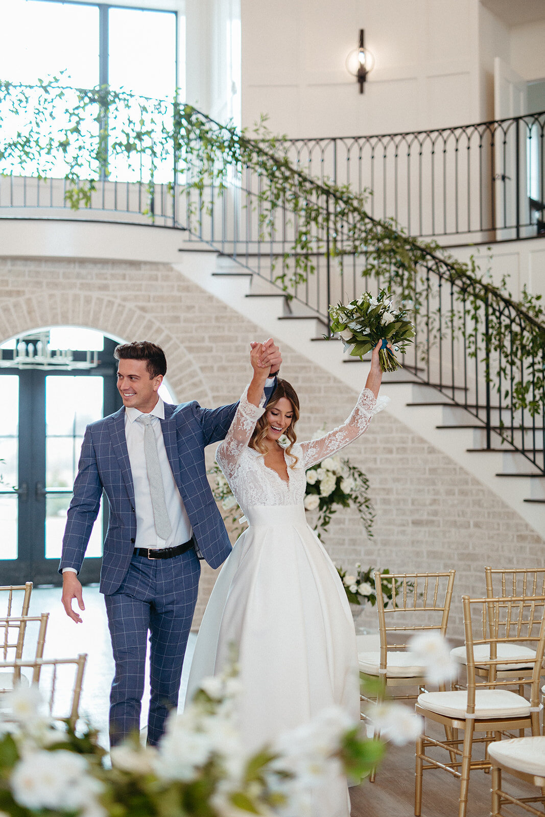 Bride and groom in a blue suit and white wedding gown walking down an aisle of gold chairs and white flowers.