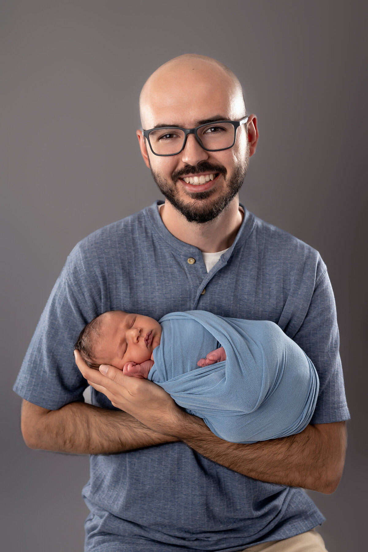 A happy father stands in a studio cradling his sleeping newborn son in his arms