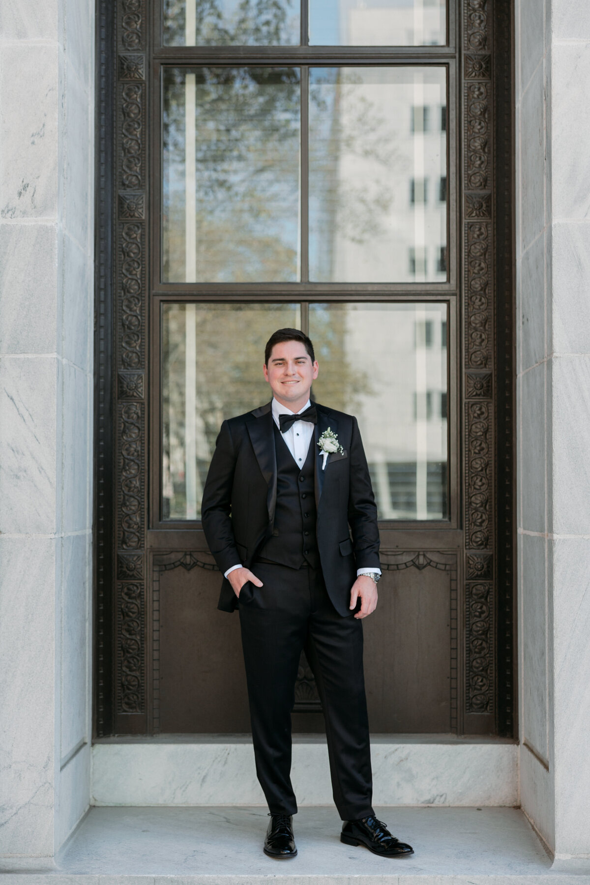 Groom standing confidently in front of a carved window frame in Philadelphia, wearing a black tuxedo.