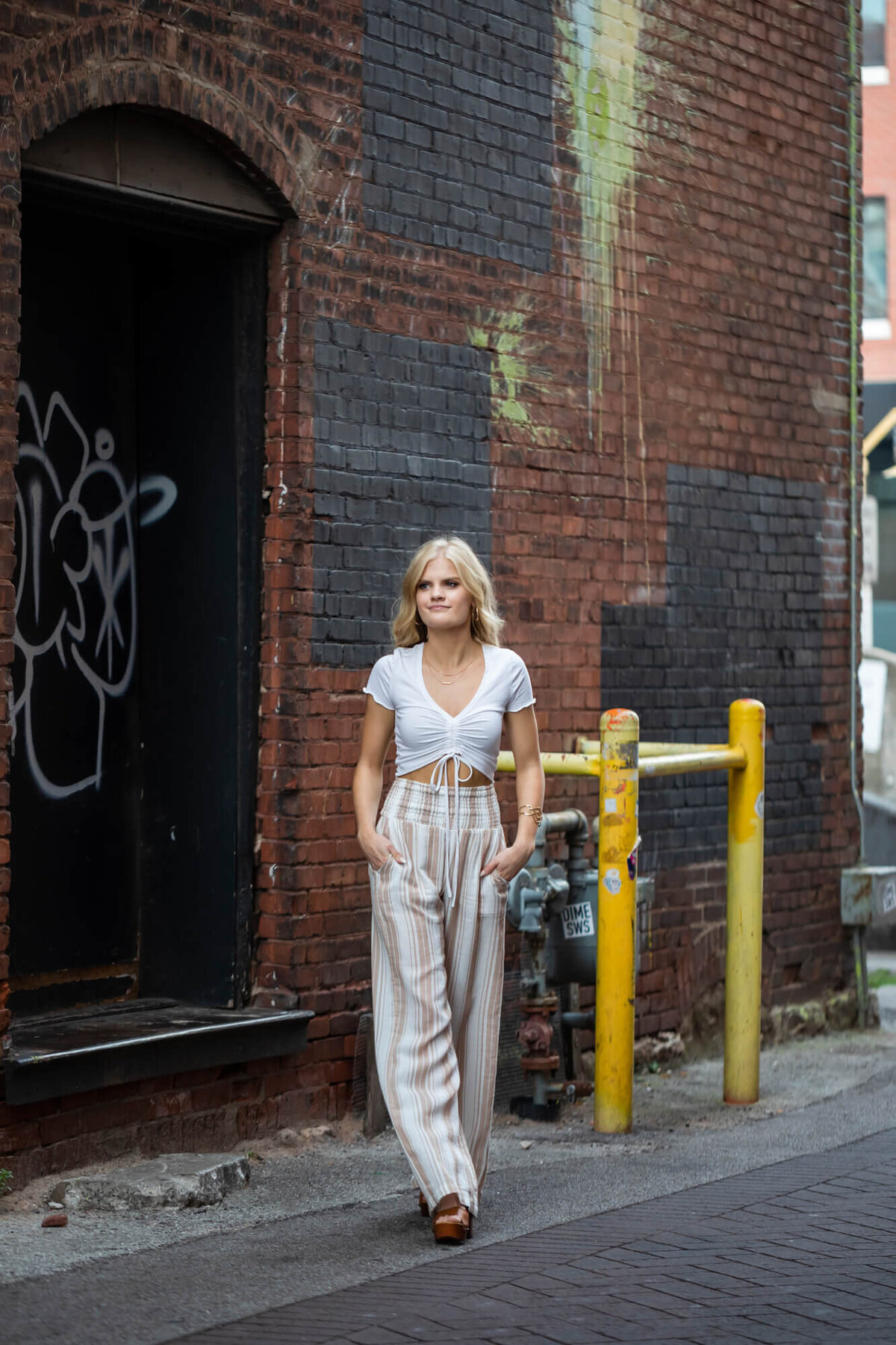 A casual senior portrait of a beautiful blonde high school senior walking in a downtown alleyway with graffiti. Captured by senior photographer Dynae Levingston.