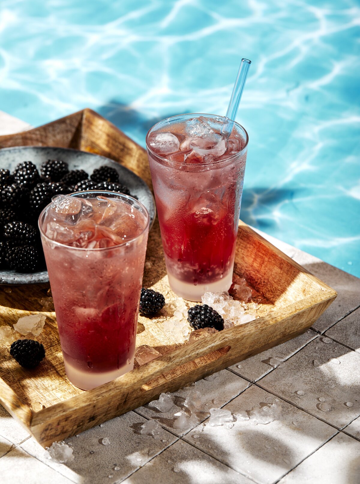 Two glasses of a berry drink with blackberries sitting next to a pool.
