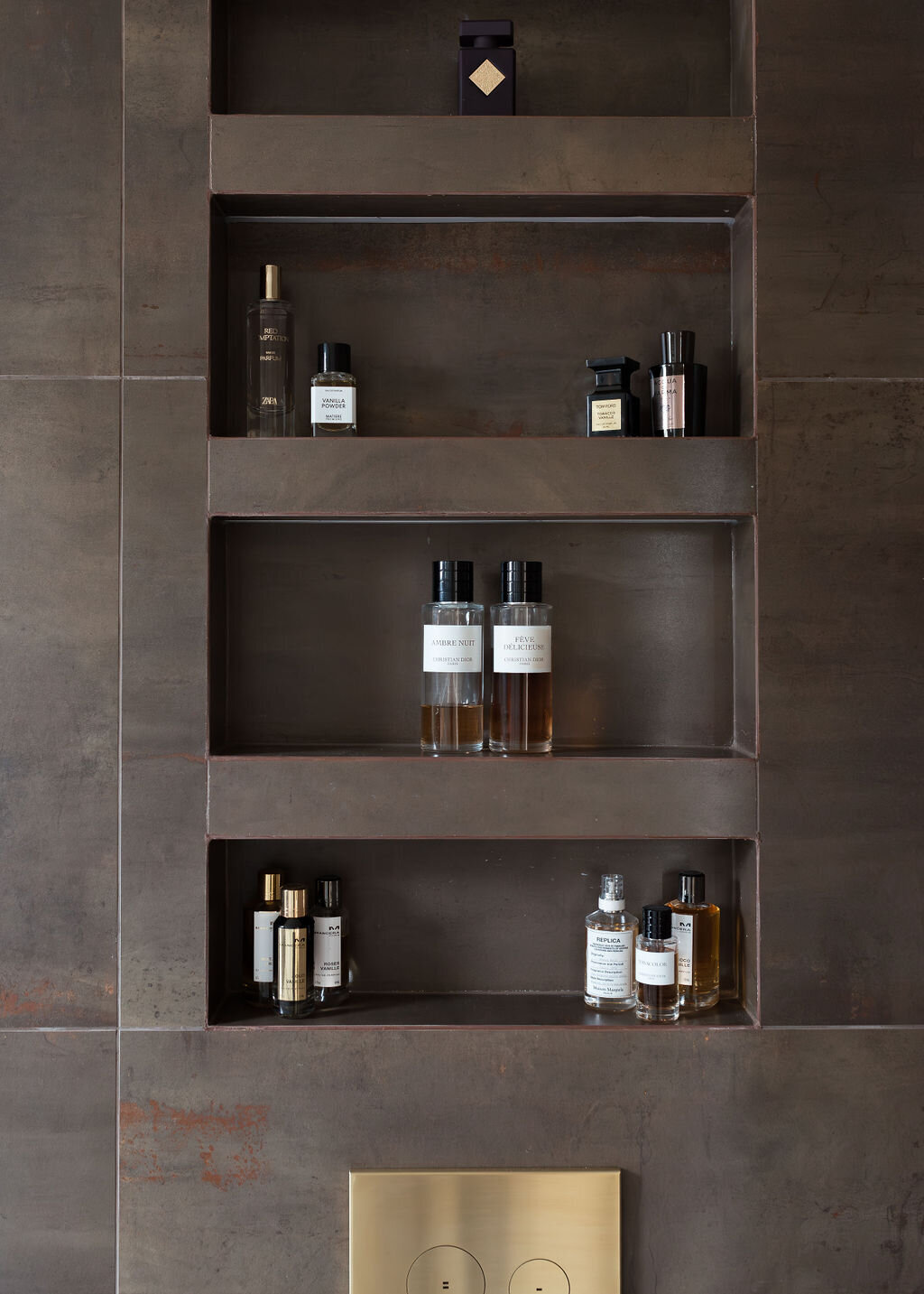 Above a toilet in a modern bathroom, shelves are built into the wall, holding toiletries. The wall has large, dark brown tiles.