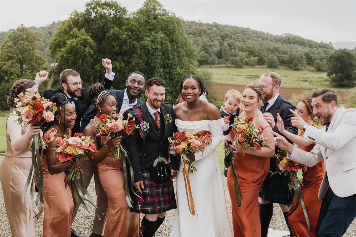 9 people, all bridesmaids and groomsmen, huddle around a bride and groom. They cheer and joyfully celebrate the couple as they pose for a photograph.