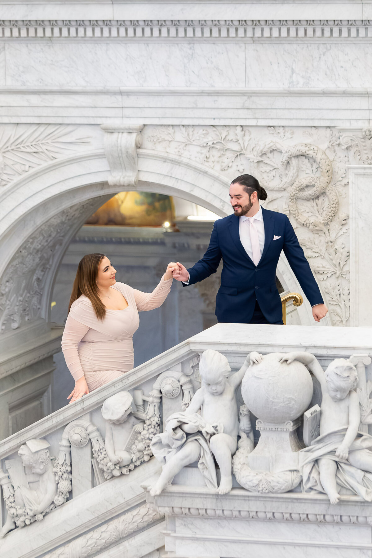 A man holds the hand of his fiancé and leads her up a set of ornate stairs in Washington D.C. He's looking back at her smiling.