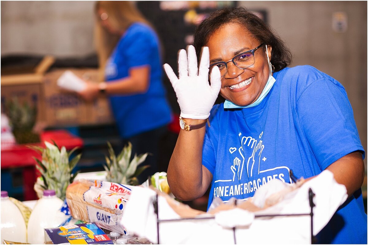a-smiling-volunteer-wearing-a-blue-shirt-and-gloves-waves-while-packing-food-at-a-community-service-event-in-indianapolis