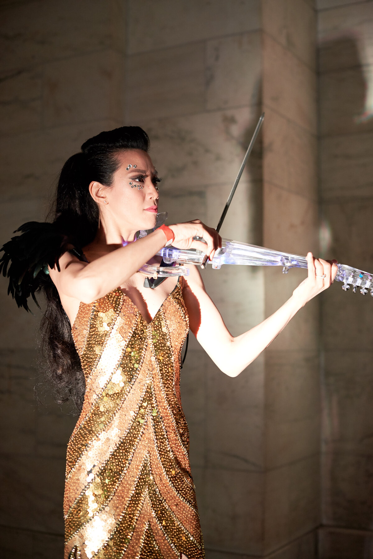 The stunning photograph was taken at the iconic New York Public Library in New York City by photographer Christian Oth. It captures a moment of sheer elegance and sophistication as a person, dressed in a sparkling gold sequin gown and adorned with a black feathered accessory, skillfully plays a clear violin. Their long, dark hair and intricate face makeup add to the glamour of the scene, set against a backdrop of a luxurious marble wall. This combination of elements creates a captivating and beautiful composition in the photograph.  Videographers: East West Films