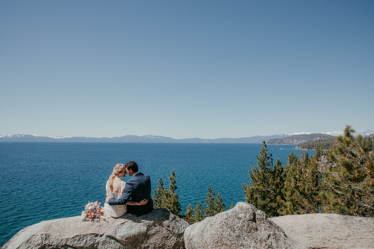 Bride and groom sitting together on a rock ledge overlooking Lake Tahoe at Logan Shoals Vista Point, with sparkling blue water and distant snow-capped peaks.