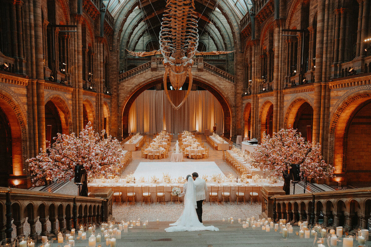 Bride and groom reviewing their wedding design in Hintze Hall at the Natural History Museum in London, with candles lining the staircase and cherry blossom trees.