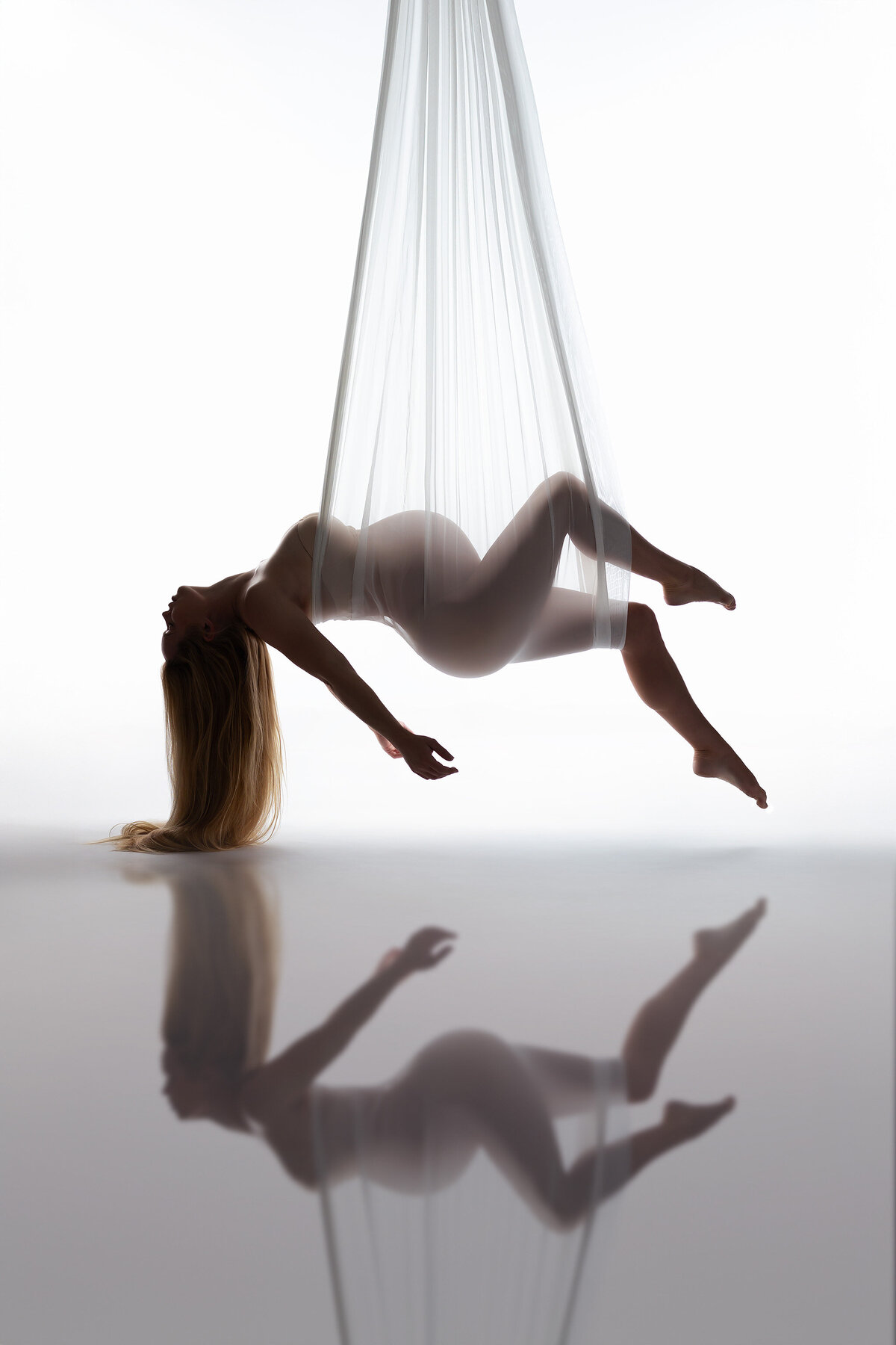 A serene silhouette of a woman gracefully suspended in sheer white fabric, her body delicately cradled and reflected on the smooth surface below. This captivating scene highlights the ethereal beauty of maternity photography in Dallas.