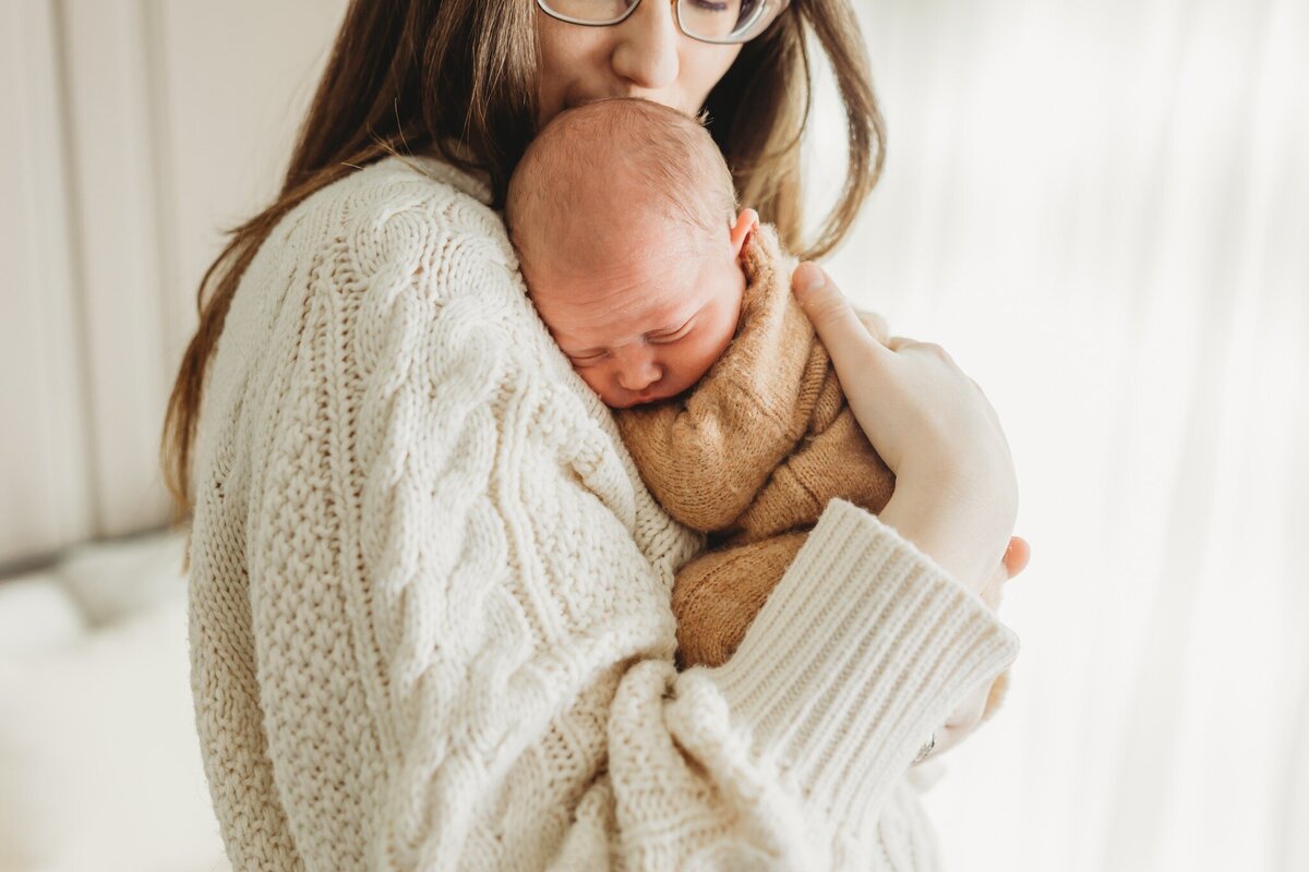 A mother wearing a chunky, white knitted sweater cradles her newborn baby close to her chest. The baby is swaddled, appearing peaceful and content. Soft natural light filters through a window in the background, creating a warm and intimate atmosphere.
